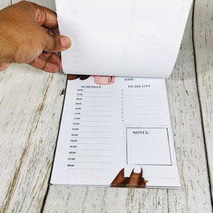 May include: A white planner with a black and white illustration of a woman's hand with a manicure. The planner has a section for a daily schedule, a to-do list, and notes.