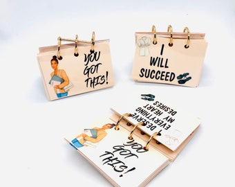 May include: Three small, beige, ring-bound notebooks with motivational quotes. The first notebook says "You got this!" and features a cartoon illustration of a woman holding a stack of papers. The second notebook says "I will succeed" and features a cartoon illustration of a woman wearing a coat. The third notebook says "You got this!" and features a cartoon illustration of a woman holding a stack of papers.