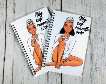 May include: Two white spiral bound notebooks with black rings. Each notebook features a black and white illustration of a woman with a headscarf holding a cup. The text "My cup runneth over" is printed on each notebook.