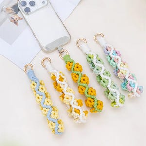 May include: Five macrame phone wrist straps featuring small knotted flower designs in blue, yellow, green, and pastel colors, each attached to a gold-toned metal ring. One strap is connected to a clear smartphone case with a triple-lens camera.