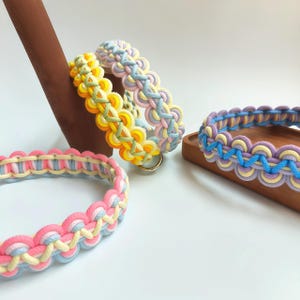 May include: Three braided rope collars featuring intricate woven patterns in pastel color palettes including pink, yellow, blue, and lavender. Each collar is constructed from multiple strands of cord with a circular metal ring attached to the side.