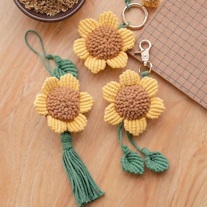 May include: Three macrame sunflower keychains made of yellow and brown cord. Two keychains feature metal lobster clasps and green leaf accents, while one includes a long green tassel. The sunflowers have textured centres and layered petals.