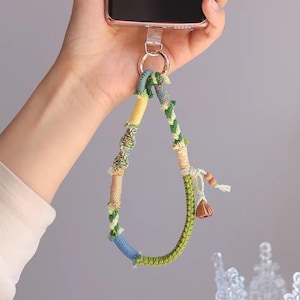 May include: A colorful macrame phone strap with a gold-tone ring. The strap features a variety of colors, including blue, green, yellow, and beige. It has a decorative tassel and wooden beads. The strap is designed to attach to a phone.