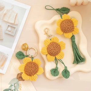 Puede incluir: Tres colgantes de girasol de macramé con centros marrones y pétalos amarillos. Un colgante tiene un llavero de metal dorado y una pequeña campana, otro un cierre de mosquetón dorado y el tercero incluye un lazo de cordón y una borla verde.