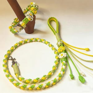 Könnte beinhalten: Ein Hundehalsband, eine Leine und ein passendes Schlüsselanhänger-Set. Das Set besteht aus geflochtenem Seil in Grün, Gelb und Weiß. Das Halsband hat eine silberne Schnalle und die Leine einen silbernen Karabiner. Das Set ist mit gänseblümchenförmigen Akzenten verziert.