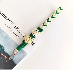 May include: A braided lanyard in green, white, and yellow, adorned with daisy-shaped embellishments. The lanyard has a gold-colored clasp and is resting on a white surface with the text "EMBRACE THE NA... I LOVE GREEN".