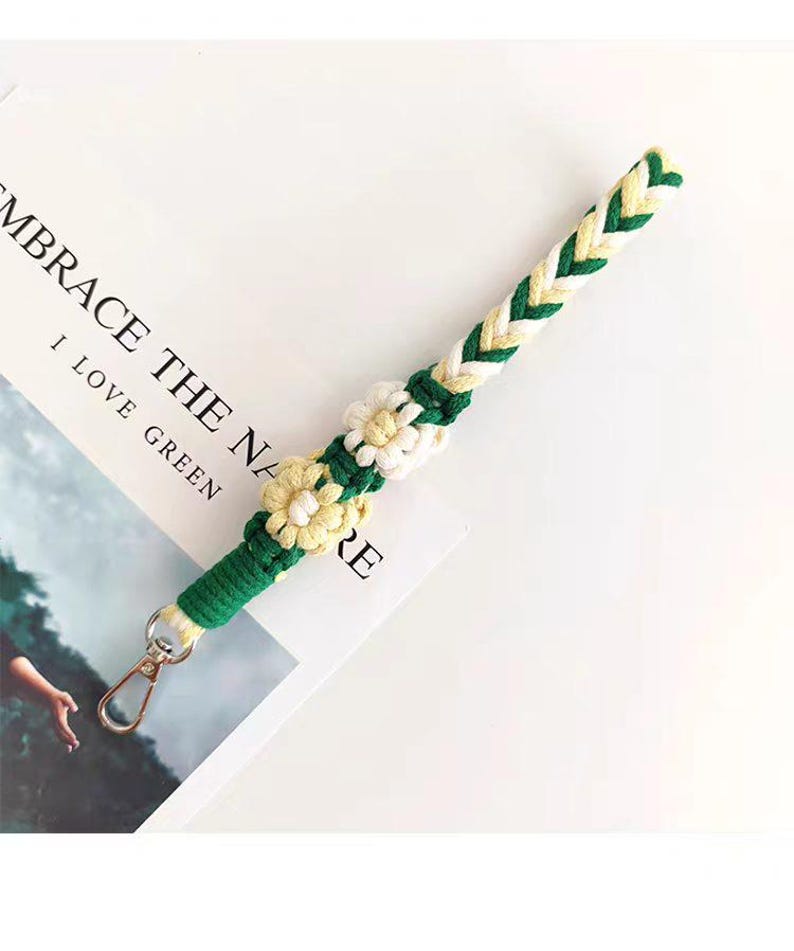 May include: A braided lanyard in green, white, and yellow, adorned with daisy-shaped embellishments. The lanyard has a gold-colored clasp and is resting on a white surface with the text "EMBRACE THE NA... I LOVE GREEN".