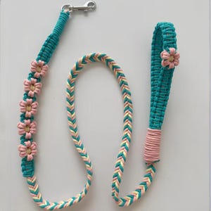 May include: A macrame dog leash crafted from teal, pale yellow, and soft pink cord. The leash features a braided design, a silver metal swivel snap hook at one end, a loop handle, and five decorative pink and yellow flower-shaped knots.