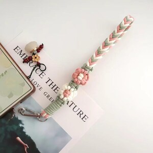 May include: A macrame phone strap in shades of pink, green, and white, adorned with floral accents. The strap features a braided design and a silver-tone clasp. The words "EMBRACE THE NATURE" are visible in the background.