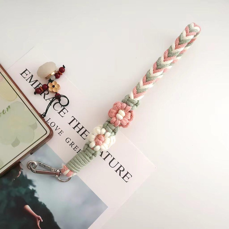 May include: A macrame phone strap in shades of pink, green, and white, adorned with floral accents. The strap features a braided design and a silver-tone clasp. The words "EMBRACE THE NATURE" are visible in the background.