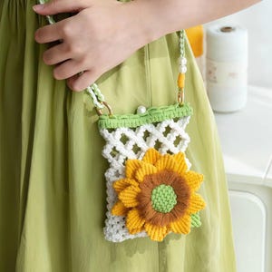 Può includere: Una borsa a tracolla in macramè bianco con una grande applicazione tridimensionale a forma di girasole giallo e marrone sul davanti. La borsa presenta una tracolla intrecciata verde chiaro con perle bianche e un bordo in macramè verde chiaro.