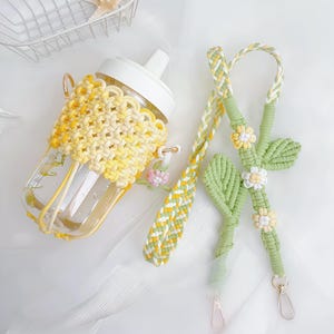 May include: A clear plastic water bottle with a white spout, encased in a yellow and cream macrame holder. A matching braided strap features green macrame leaves and small white and yellow flower accents, finished with gold-toned metal clasps.