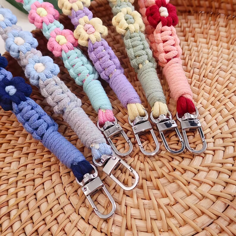 May include: Colorful macrame bag straps with flower accents. Each strap features a different color scheme, including blue, pink, yellow, and purple, with silver-tone metal clasps for easy attachment. The straps are displayed on a woven surface.