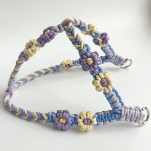 May include: A macrame pet harness crafted from braided cord in shades of lavender, light blue, and pale yellow. The design features integrated flower-shaped knots along the straps and includes two silver-toned metal D-rings for attachment points.