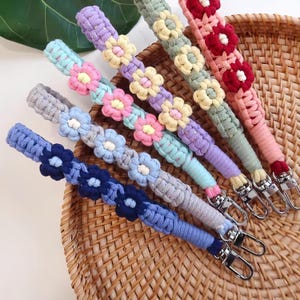 Peut inclure: Plusieurs bandoulières de sac en macramé colorées. Chaque sangle présente un schéma de couleurs différent, notamment bleu, gris, rose, jaune et vert, avec des motifs floraux. Les sangles sont équipées de fermoirs argentés.