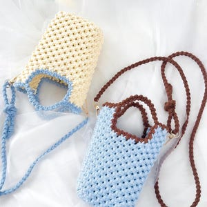 Könnte beinhalten: Zwei kleine Häkeltaschen mit offenem Webmuster. Eine Tasche ist cremefarben mit hellblauem Rand und passendem hellblau geflochtenem Riemen. Die zweite Tasche ist hellblau mit dunkelbraunem Rand und passendem dunkelbraun geflochtenem Riemen.