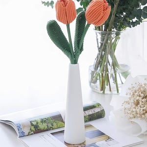 May include: Two orange crocheted tulip flowers with textured green stems and leaves, displayed in a tall, slender, matte white ceramic vase. The tulips feature a ribbed, yarn-based construction, standing upright with distinct petals and long, pointed foliage.