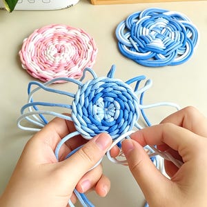 Può includere: Tre sottobicchieri circolari intrecciati con corda. Un sottobicchiere presenta un motivo a spirale rosa e bianco, un altro mostra un complesso design a nodo blu e bianco, e il terzo è una spirale blu e bianca intrecciata a mano.