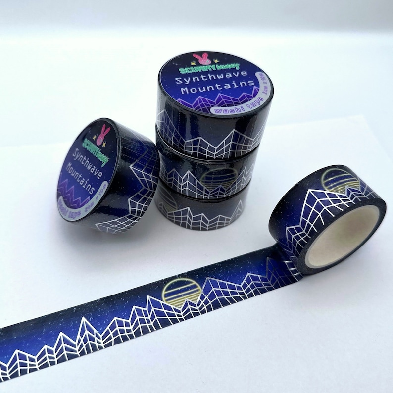 Op de afbeelding: Een rol washi tape met een blauw en wit geometrisch bergontwerp en een gouden zonsondergang. De tape is gelabeld "Synthwave Mountains" en heeft een cartoon konijnenlogo.