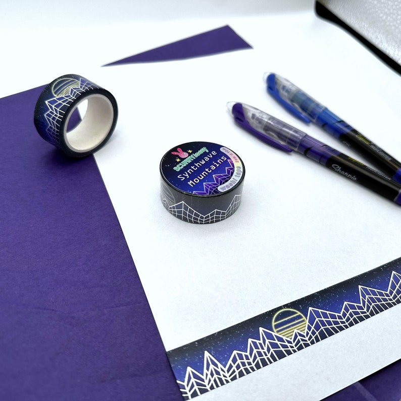 Op de afbeelding: Twee rollen washi tape met een zwarte achtergrond en witte geometrische ontwerpen. Een rol heeft de tekst "Synthwave Mountains" en een roze konijn met een ster. De andere rol heeft een witte maan en sterren.