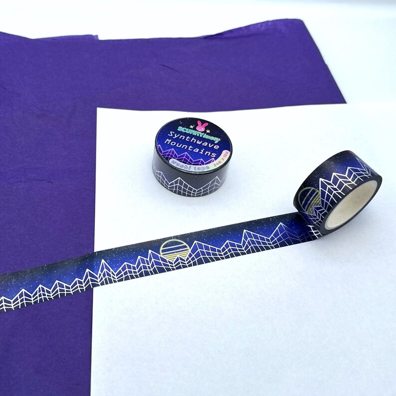 Op de afbeelding: Een rol washi tape met een zwarte achtergrond en gouden accenten. De tape heeft een herhalend patroon van bergen, sterren en een zon. De tape is gelabeld "Synthwave Mountains" en is verpakt in een doorzichtige plastic container.