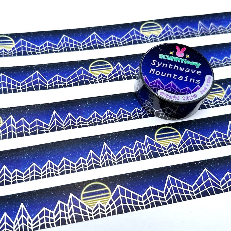 Op de afbeelding: Een rol washi tape met een blauw en wit geometrisch bergontwerp en een gele zon. De tape is gelabeld met "Synthwave Mountains" en "washi tape" met een konijnenlogo.