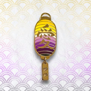Puede incluir: Pin de color dorado con forma de linterna japonesa de papel con la palabra "amor" en caracteres japoneses y un pequeño amuleto con dos corazones colgando de la parte inferior.