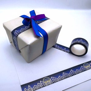 Op de afbeelding: Een witte cadeaudoos ingepakt met blauw en paars lint en versierd met een zwart-wit geometrisch patroon washi tape. De tape heeft een herhalend ontwerp van bergen en sterren.