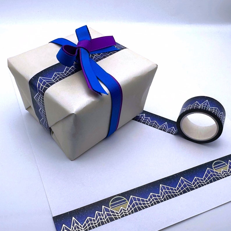 Op de afbeelding: Een witte cadeaudoos ingepakt met blauw en paars lint en versierd met een zwart-wit geometrisch patroon washi tape. De tape heeft een herhalend ontwerp van bergen en sterren.