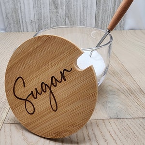 May include: A round glass sugar bowl with a bamboo lid. The lid has a cutout for a spoon and the word "Sugar" is engraved on the lid.