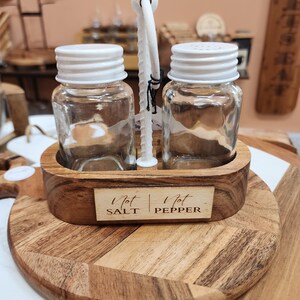 May include: Wooden salt and pepper shaker holder with a metal stand. The holder has two glass shakers with white lids and the text "Not Salt" and "Not Pepper" on the front.