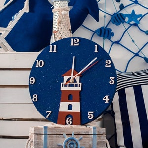 Puede incluir: Un reloj azul y blanco con un diseño de faro. El reloj tiene un fondo blanco con números azules y un faro rojo en el centro. El reloj está rodeado de decoración náutica, incluyendo una almohada a rayas azules y blancas y una red de pesca.