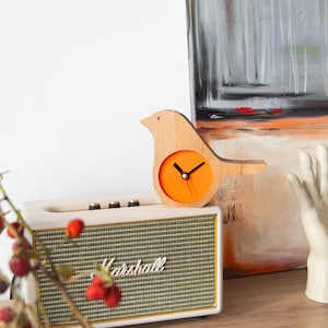 May include: A wooden bird-shaped clock with an orange face and black hands. The clock is sitting on top of a white speaker with the word "Marshall" printed on the front. There is a white hand sculpture in the background.