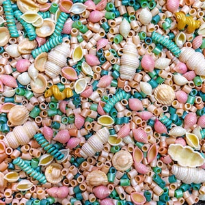 May include: A collection of assorted dried pasta shapes including shells, tubes, and spirals in a mix of colors such as light pink, teal, beige, and gold. The various pasta pieces are scattered together to create a dense, textured surface.
