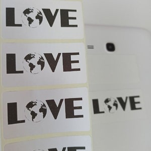 Puede incluir: Un rollo de pegatinas rectangulares blancas con la palabra "LOVE" en gris oscuro, cada una con un gráfico de globo terráqueo. Varias pegatinas son visibles en el rollo. El fondo es una superficie blanca.