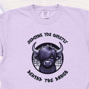 May include: Light purple t-shirt featuring a cartoon Highland cow illustration. The cow has long, dark hair and is inside a circular frame with the text "Judging You Quietly Behind The Bangs". The shirt is a soft, comfortable material.