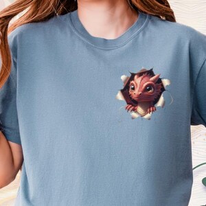 May include: A light blue t-shirt featuring a detailed illustration of a baby dragon bursting through a torn hole. The dragon has red and brown scales, large eyes, and a curious expression. The shirt is made of soft material.