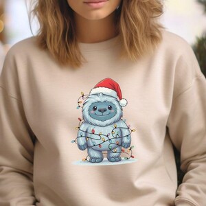 May include: A beige sweatshirt featuring a cartoon yeti wearing a Santa hat and wrapped in Christmas lights. The yeti is light blue with a friendly expression. A red Christmas ornament is visible in the background.