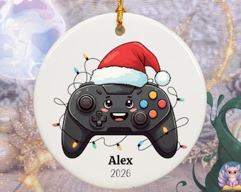 May include: A round white ceramic ornament featuring a cartoon black video game controller wearing a red Santa hat. The controller is wrapped in colorful Christmas lights and has a smiling face. The text Alex and 2026 is printed in black below.