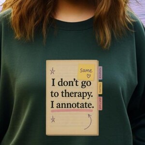 May include: Dark green sweatshirt with the text "I don't go to therapy. I annotate." printed on a book graphic. The graphic includes a yellow sticky note that says "Same" with a heart symbol. The sweatshirt is a solid color.