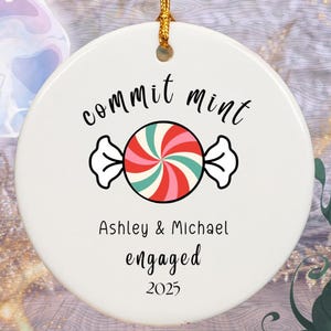 May include: White ceramic ornament with a gold string, featuring the words "commit mint" above a candy graphic. Below the candy, it reads "Ashley & Michael engaged 2025".