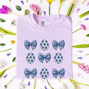 May include: A lavender t-shirt with a pattern of blue bows and blue and white eggs. The shirt has a tag that says "Comfort Colors" on it.