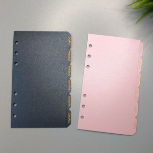 May include: Two planner divider sheets with six ring holes on the left side. One sheet is dark gray with gold text reading GOALS, PROJECT, INCOME, EXPENSES, TRACKER, SOCIALS. The other is light pink with gold text reading BUDGET, INCOME, EXPENSES, WISHLIST, GOALS, NOTES.