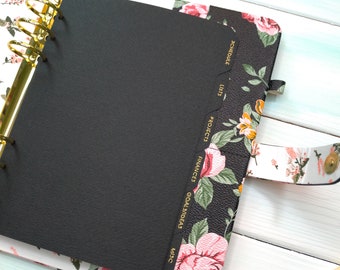 May include: A personal planner with a black floral cover featuring pink roses and green leaves. It includes a gold-toned metal ring binder mechanism and black tabbed dividers labeled in gold foil with Schedule, Lists, Projects, Finances, Goals/Ideas, and Misc.