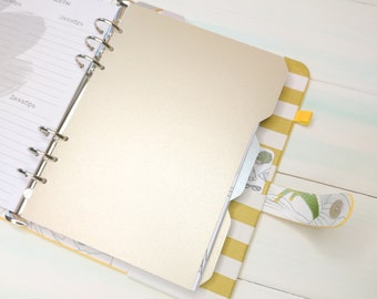 Op de afbeelding: Een ringbandplanner met een metallic gouden tabblad, een geel-wit gestreepte stoffen kaft en een sluiting met drukknoop. De binnenpagina's bevatten gedrukte tekst in het Cyrillisch, waaronder de maanden oktober en december.