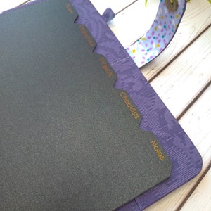 May include: A purple planner with a textured cover and a magnetic strap featuring a colorful confetti pattern. Inside, dark gray tabbed dividers are labeled in orange text with the categories: Index, Objectives, Projects, Checklists, and Notes.