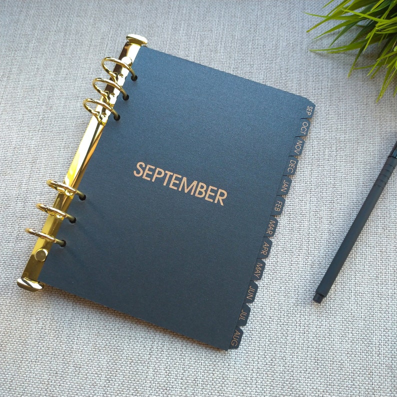 May include: A black planner with gold-colored metal rings and the word SEPTEMBER printed in gold on the front cover. The planner features side tabs labeled with months from September through August, and a black pen is positioned to the right.