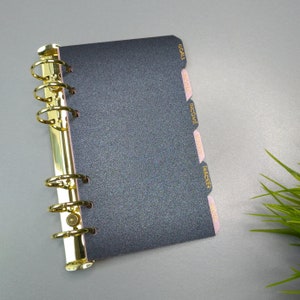 May include: A dark gray glittery personal planner with a gold-toned six-ring metal binder mechanism. The planner features side tabs labeled in gold text with the words GOALS, INCOME, INCOME, MISC, TRACKER, and NOTES on light pink dividers.
