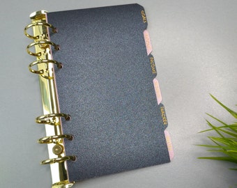 May include: A dark gray glittery personal planner with a gold-toned six-ring metal binder mechanism. The planner features side tabs labeled in gold text with the words GOALS, INCOME, INCOME, MISC, TRACKER, and NOTES on light pink dividers.