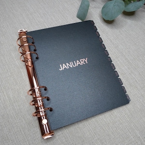 May include: A black planner with a rose gold metal ring binder mechanism on the left side. The cover features the word JANUARY in rose gold foil lettering. The right edge has twelve black index tabs labeled with the months from Jan to Dec.